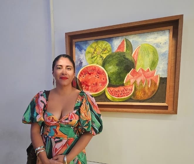Woman in a colorful floral dress stands beside Frida Kahlo's watermelon still life painting.