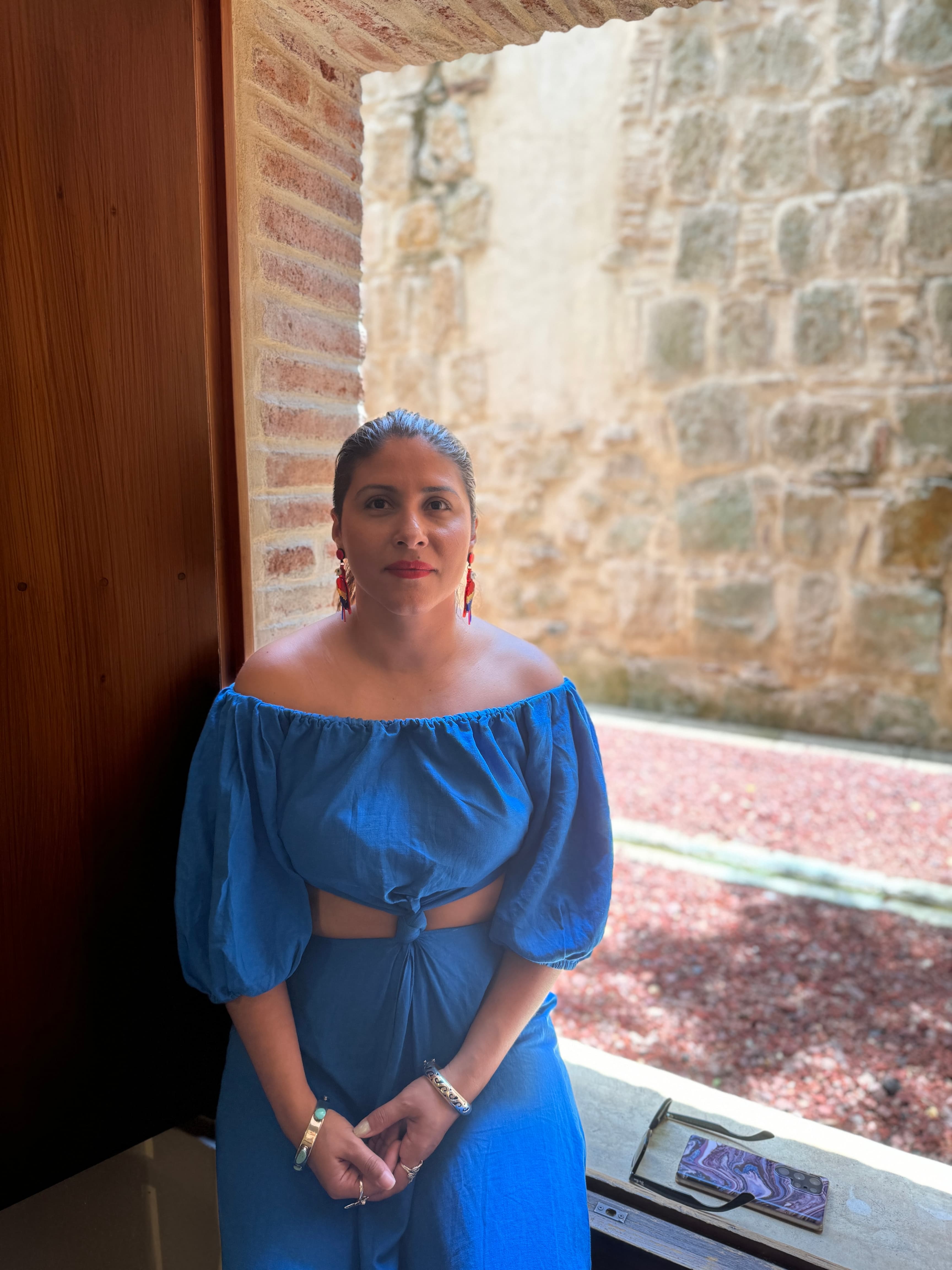 Woman in a blue off-the-shoulder outfit sits in a rustic stone window frame.